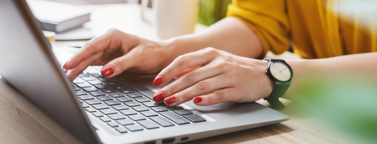 A woman in a yellow ochre shirt and red nails is typing on a laptop with a coffee to her right. This image depicts someone working on improving SEO.