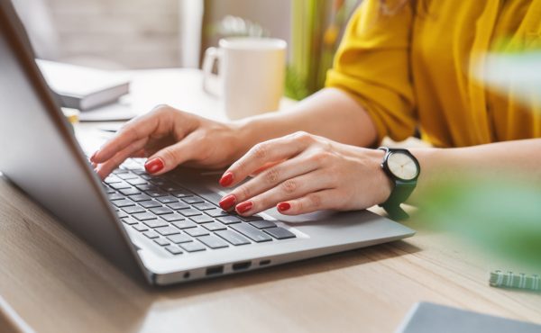 A woman in a yellow ochre shirt and red nails is typing on a laptop with a coffee to her right. This image depicts someone working on improving SEO.
