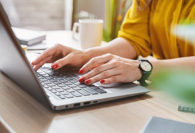 A woman in a yellow ochre shirt and red nails is typing on a laptop with a coffee to her right. This image depicts someone working on improving SEO.