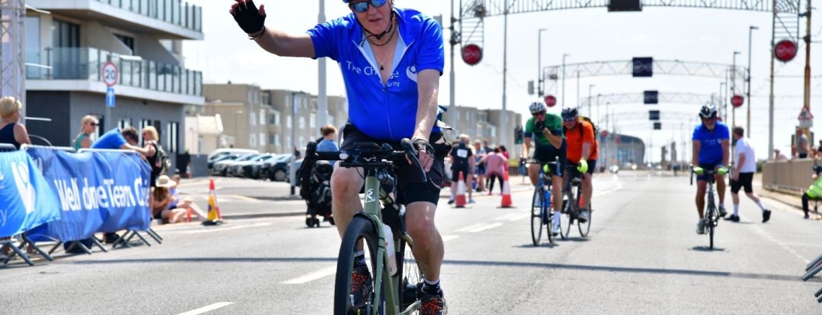 Paul Barlow, director of Smith Goodfellow, is riding his bike across the finish line in his blue The Christie charity vest. He is wearing a helmet and has his hand raised in celebration.