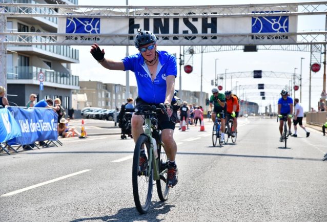 Paul Barlow, director of Smith Goodfellow, is riding his bike across the finish line in his blue The Christie charity vest. He is wearing a helmet and has his hand raised in celebration.