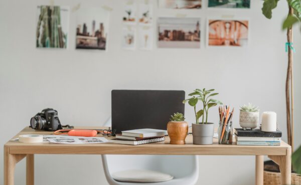 the image is of a light wooden home office desk. It has some bright accessories, such as plant pots and stationery, in addition to an open laptop and camera. There is a selection of prints on the wall which are out of focus.