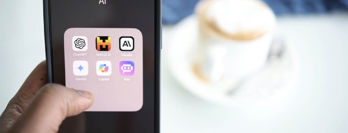A person is holding a mobile phone with the screen visible. It has a folder open that is labelled 'AI' and contains icons of AI apps: ChatGPT, Mistral AI, Claude, Gemini, Copilot, and Poe. There is a pale table and a white cup and saucer with coffee in the background.