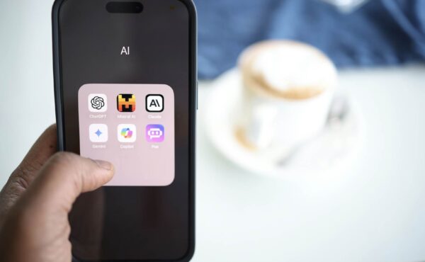 A person is holding a mobile phone with the screen visible. It has a folder open that is labelled 'AI' and contains icons of AI apps: ChatGPT, Mistral AI, Claude, Gemini, Copilot, and Poe. There is a pale table and a white cup and saucer with coffee in the background.