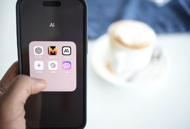 A person is holding a mobile phone with the screen visible. It has a folder open that is labelled 'AI' and contains icons of AI apps: ChatGPT, Mistral AI, Claude, Gemini, Copilot, and Poe. There is a pale table and a white cup and saucer with coffee in the background.