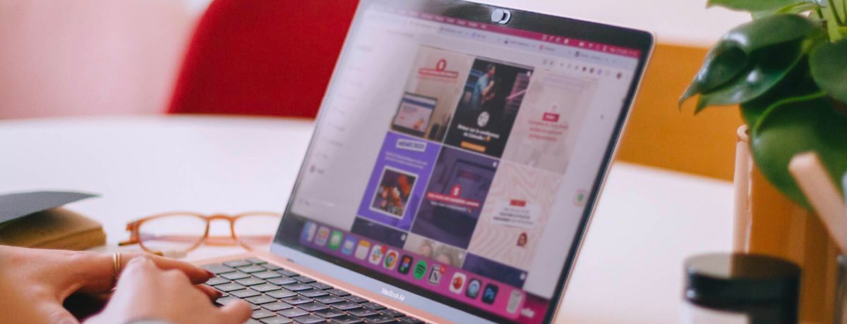 There is a rose-coloured MacBook Air laptop on a table at an angle. Hands are poised ready to type. There are glasses on the table to the left of the laptop and a candle and leafy plant to the right. On the screen is an Instagram grid layout of branded social media graphics.