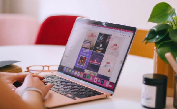 There is a rose-coloured MacBook Air laptop on a table at an angle. Hands are poised ready to type. There are glasses on the table to the left of the laptop and a candle and leafy plant to the right. On the screen is an Instagram grid layout of branded social media graphics.