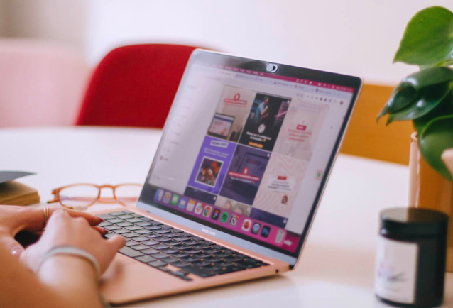 There is a rose-coloured MacBook Air laptop on a table at an angle. Hands are poised ready to type. There are glasses on the table to the left of the laptop and a candle and leafy plant to the right. On the screen is an Instagram grid layout of branded social media graphics.
