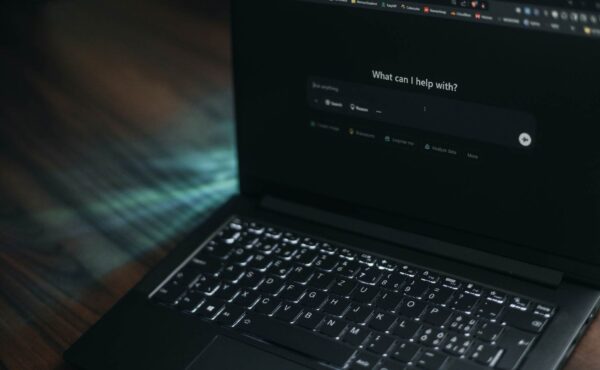 A dark image showing a laptop at an angle with the screen showing an AI chat bot landing page. The ChatGPT dashboard reads: "what can I help with?" and the cursor is ready in the prompt box.