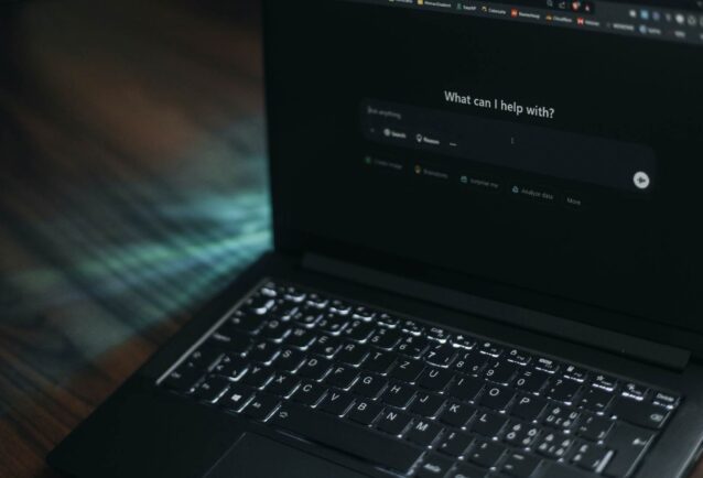 A dark image showing a laptop at an angle with the screen showing an AI chat bot landing page. The ChatGPT dashboard reads: "what can I help with?" and the cursor is ready in the prompt box.