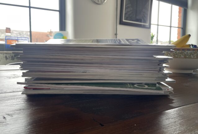 A stack of construction industry magazines are placed on a dark wood meeting room table with the fore edge facing towards the camera. In the background, items that are just visible: two big windows, pictures on the wall, a clock, and awards and accreditations on the windowsill.