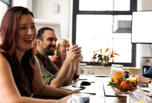Meeting room with members of the team smiling and laughing. Wooden table has a bowl of fruit and mugs.